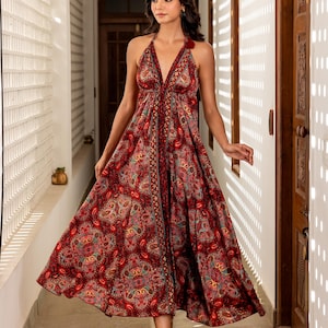May include: A halter-neck maxi dress with a vibrant paisley pattern in shades of red, pink, and gold. The dress features a flowing skirt and a V-neckline. The model is wearing beige sandals. The dress is suitable for a special occasion.