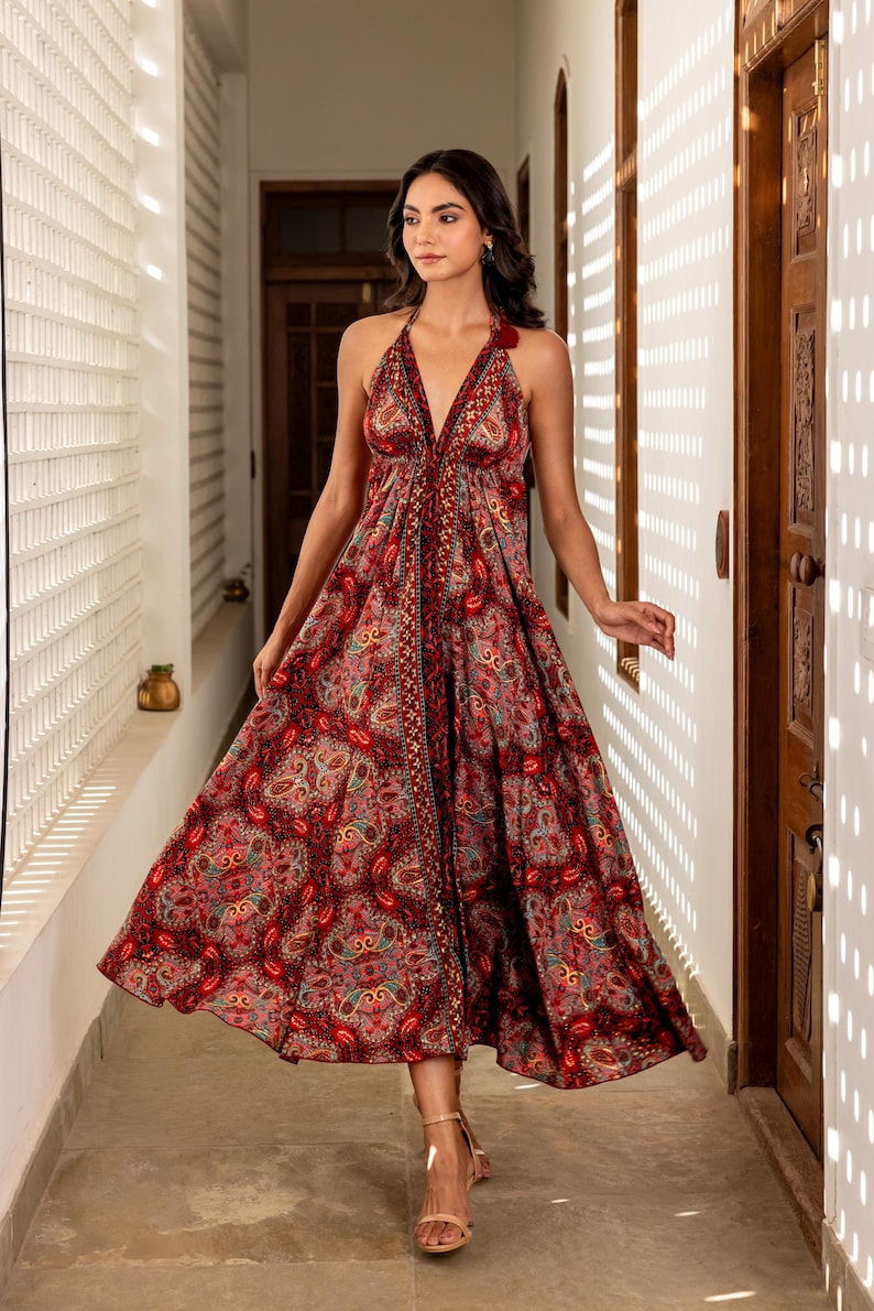 May include: A halter-neck maxi dress with a vibrant paisley pattern in shades of red, pink, and gold. The dress features a flowing skirt and a V-neckline. The model is wearing beige sandals. The dress is suitable for a special occasion.