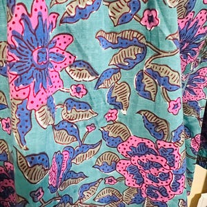 May include: Close-up of a fabric with a floral pattern. The design features large flowers in shades of pink and blue, set against a teal background. The leaves are a mix of gray and blue, creating a vibrant and detailed textile.