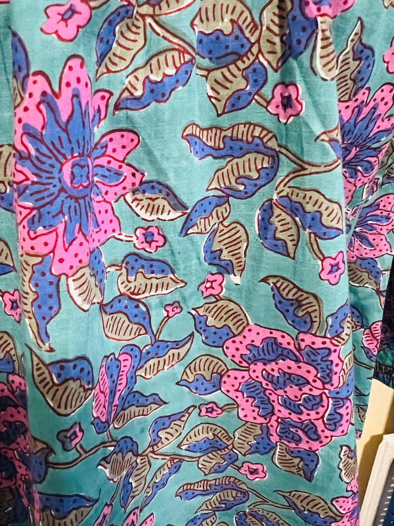 May include: Close-up of a fabric with a floral pattern. The design features large flowers in shades of pink and blue, set against a teal background. The leaves are a mix of gray and blue, creating a vibrant and detailed textile.