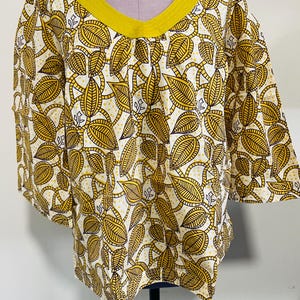 May include: A white and yellow leaf-patterned top with a yellow V-neckline and short sleeves. The fabric features a repeating design of stylized leaves and vines in yellow and brown against a white background. The top is displayed on a mannequin.