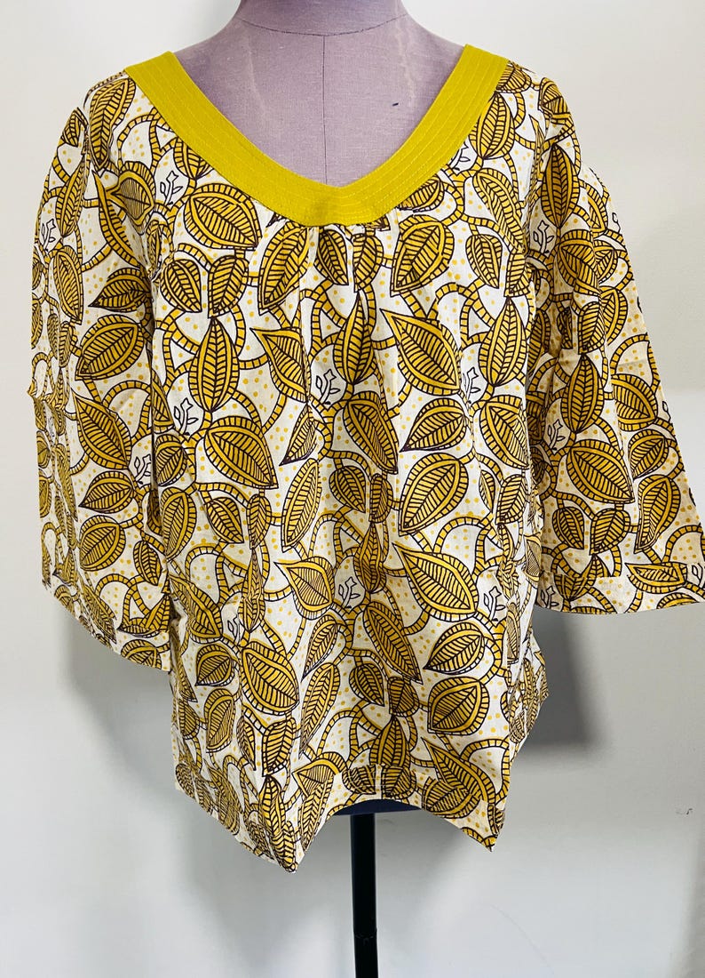 May include: A white and yellow leaf-patterned top with a yellow V-neckline and short sleeves. The fabric features a repeating design of stylized leaves and vines in yellow and brown against a white background. The top is displayed on a mannequin.