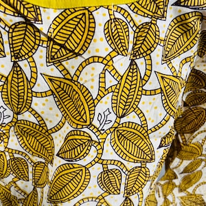 May include: Close-up of a white garment with a repeating pattern of yellow leaves and vines. The leaves have brown outlines and are filled with yellow lines. The garment has a yellow neckline.