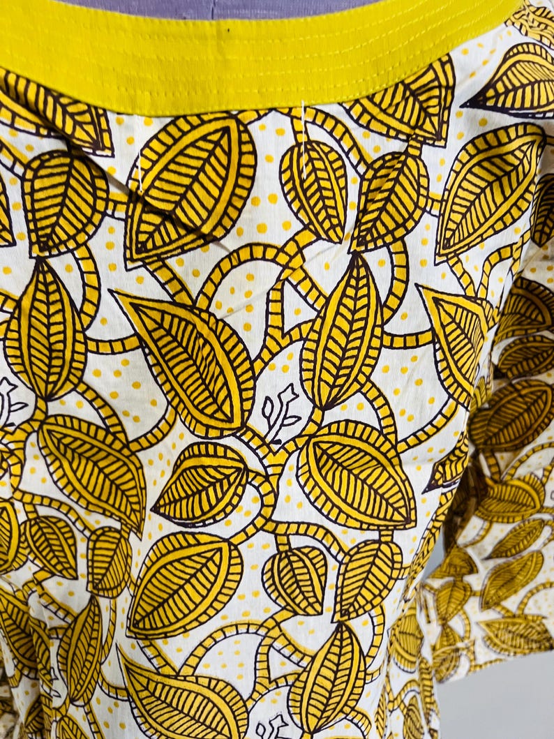 May include: Close-up of a white garment with a repeating pattern of yellow leaves and vines. The leaves have brown outlines and are filled with yellow lines. The garment has a yellow neckline.