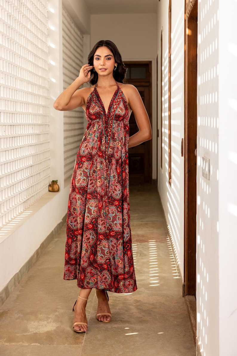 May include: A red paisley print halter dress with a V-neckline and a flowing skirt. The dress features a detailed pattern and is worn with neutral-toned heels. The dress is a midi length and is suitable for a variety of occasions.