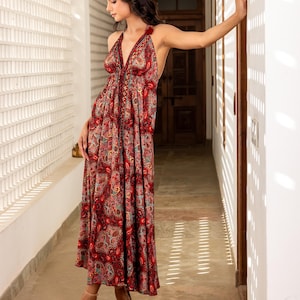 May include: A long, halter-neck dress with a paisley pattern in shades of red, pink, and green. The dress has a flowing silhouette and is worn with neutral-colored sandals. The dress is suitable for a variety of occasions.