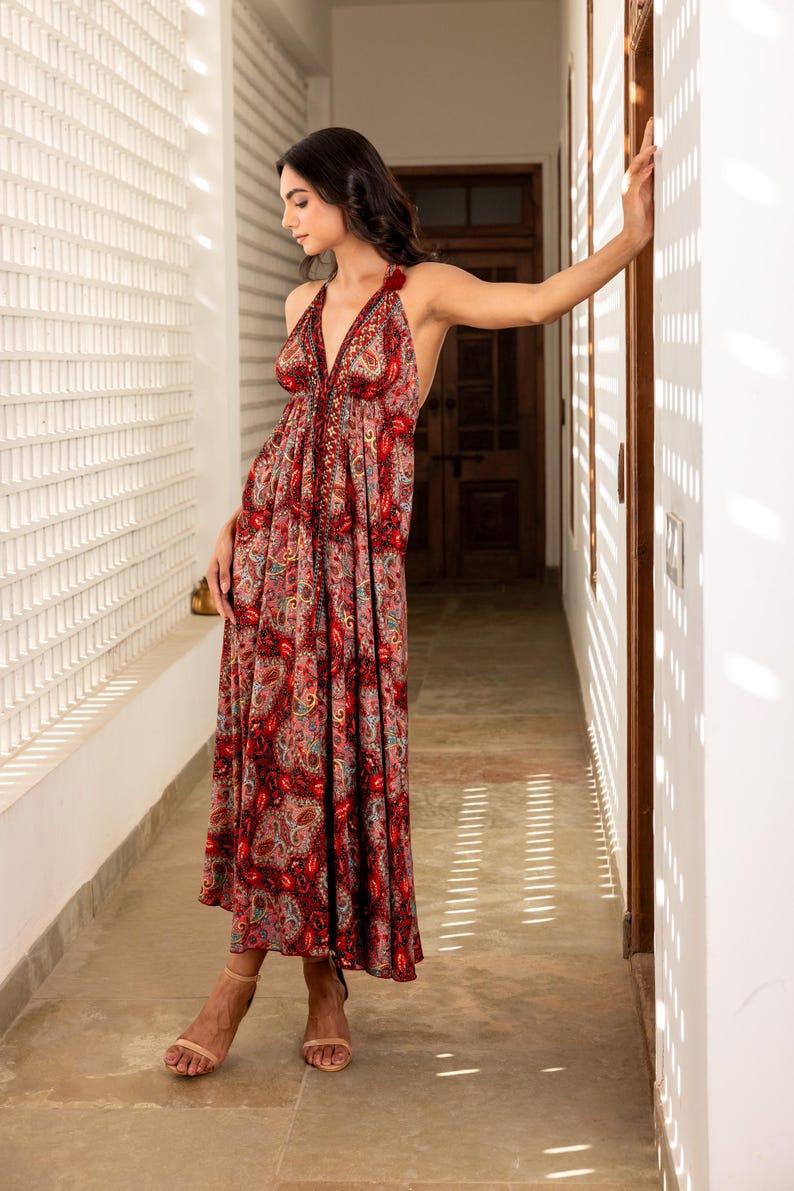 May include: A long, halter-neck dress with a paisley pattern in shades of red, pink, and green. The dress has a flowing silhouette and is worn with neutral-colored sandals. The dress is suitable for a variety of occasions.