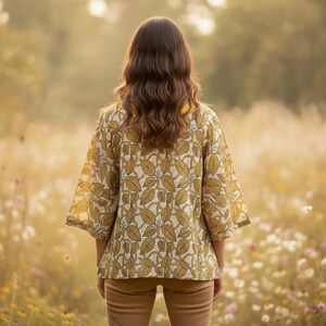 May include: A woman wearing a yellow and cream-colored kimono-style top with a leaf pattern. The top has three-quarter sleeves and a yellow collar. She is standing in a field of tall grass and wildflowers, wearing tan pants.