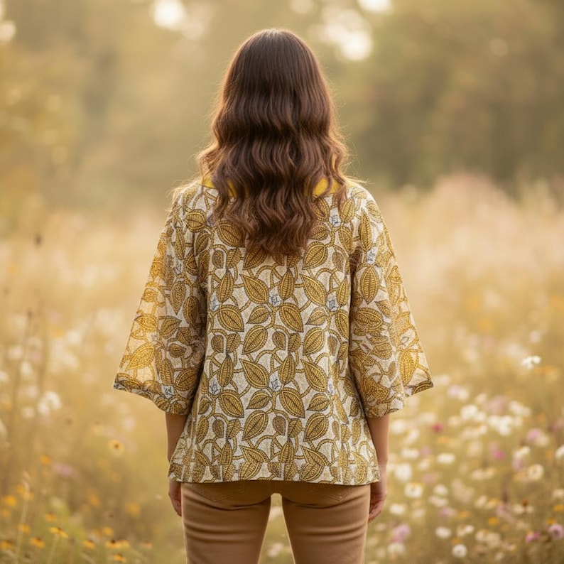 May include: A woman wearing a yellow and cream-colored kimono-style top with a leaf pattern. The top has three-quarter sleeves and a yellow collar. She is standing in a field of tall grass and wildflowers, wearing tan pants.