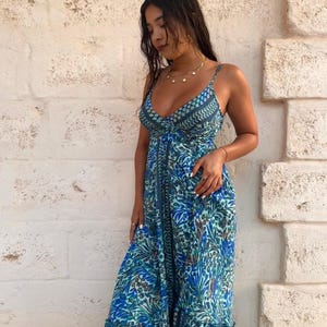 May include: A long, flowing dress with a blue and teal floral pattern. The dress has spaghetti straps and a V-neck. The fabric looks lightweight, with a tiered skirt. The model is standing in front of a textured, light-coloured wall.