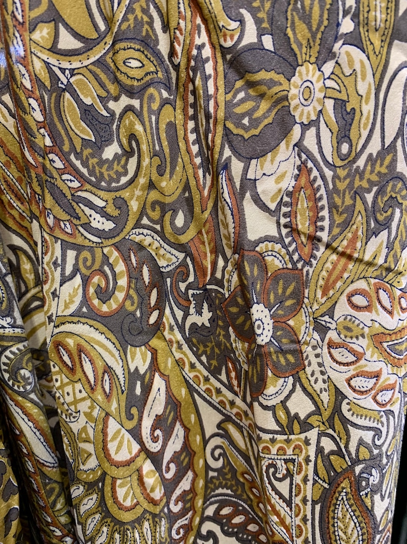 May include: Close-up of a fabric with a paisley pattern in shades of brown, olive green, and cream. The design features floral and teardrop shapes, creating a vintage or bohemian aesthetic. The fabric appears to be soft and flowing.