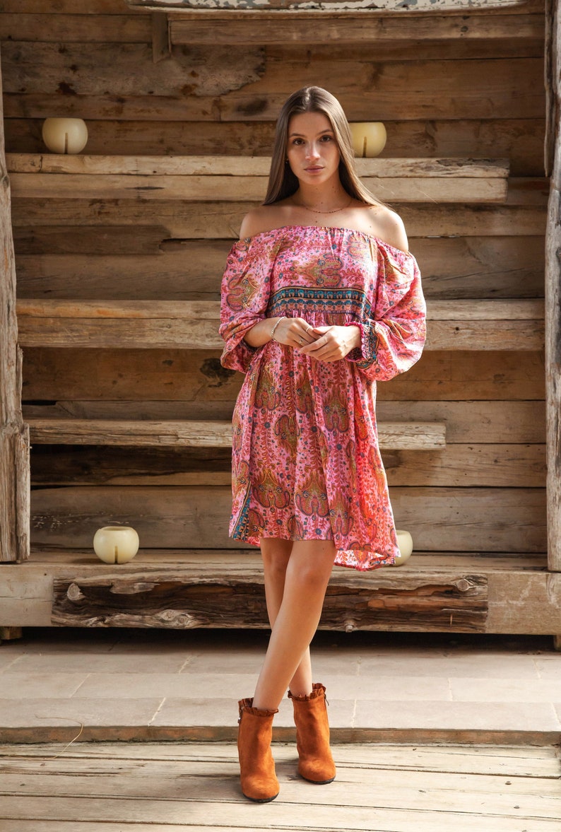 Puede incluir: Vestido rosa con hombros descubiertos y estampado de cachemira, con mangas largas y dobladillo corto. El vestido se combina con botines de ante marrones. El fondo es una estructura de madera.