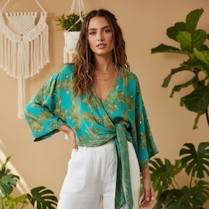 May include: A teal wrap top with a gold geometric pattern and a tie closure. The top has long sleeves and a V-neckline. The model is wearing white trousers. The background is a neutral colour.