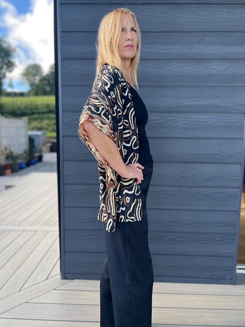 May include: A black top and wide-leg pants are paired with a patterned kimono. The kimono features a black background with a cream and brown abstract design. The outfit is worn outdoors.