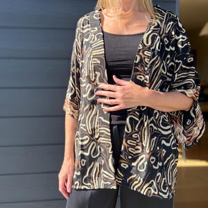May include: A black kimono-style jacket with a cream and brown abstract pattern. The jacket has wide sleeves and is open in the front. The model is wearing a black top and black pants. The background is a gray wall.