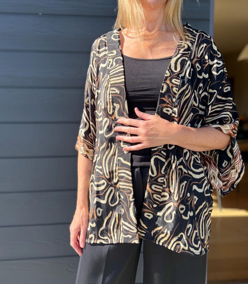 May include: A black kimono-style jacket with a cream and brown abstract pattern. The jacket has wide sleeves and is open in the front. The model is wearing a black top and black pants. The background is a gray wall.