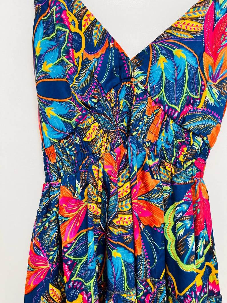 May include: A vibrant, sleeveless dress with a V-neckline. The fabric features a bold floral pattern in shades of blue, orange, pink, and yellow against a dark blue background. The dress has a gathered waist and a flowing skirt.