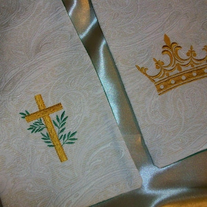 May include: Ivory-colored religious stole with gold embroidered cross and crown designs. The stole has a paisley pattern and a green border. The cross is adorned with green leaves.