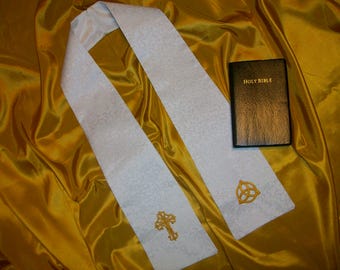 Op de afbeelding: Witte stola van stof met gouden borduurwerk van een kruis en een Keltische knoop. De stola ligt op een gouden stofoppervlak met een zwart boek waarop "HOLY BIBLE" staat.