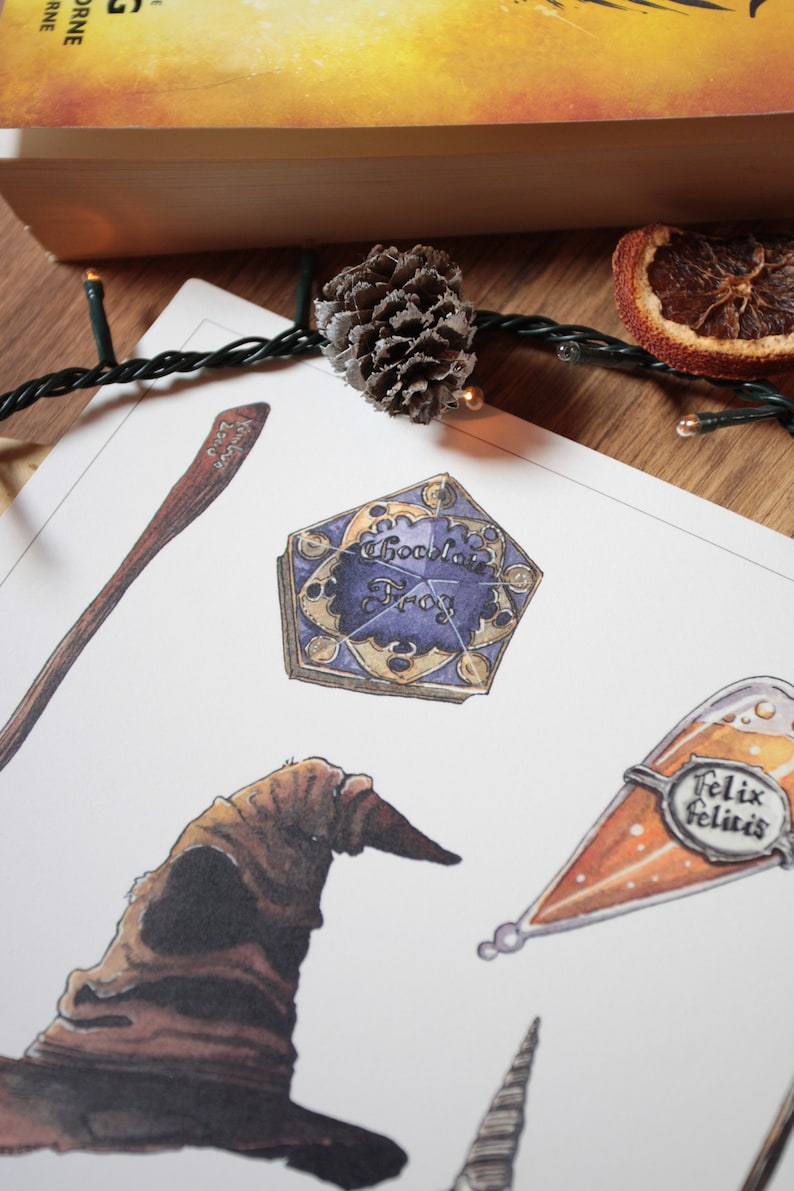 Op de afbeelding: Een witte kaart met illustraties van verschillende Harry Potter-thema-items, waaronder een chocolade kikker doos, een Sorteerhoed, een toverstaf, een fles Felix Felicis en een gouden snitch. De kaart ligt op een houten oppervlak met een dennenappel en een gedroogde sinaasappelschijf.
