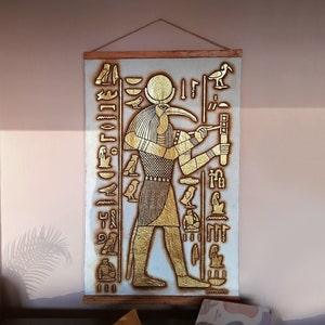 May include: A gold-colored wall hanging with an Egyptian god depicted in a standing position. The god has a bird's head with a long beak and is holding a staff. The background is white with hieroglyphs in gold.