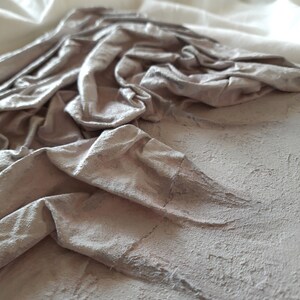 K&ouml;nnte beinhalten: Nahaufnahme einer strukturierten, hellgrauen Oberfl&auml;che mit einem zerknitterten Stoffst&uuml;ck darauf. Der Stoff ist hellgrau und hat eine weiche, knitterige Textur.