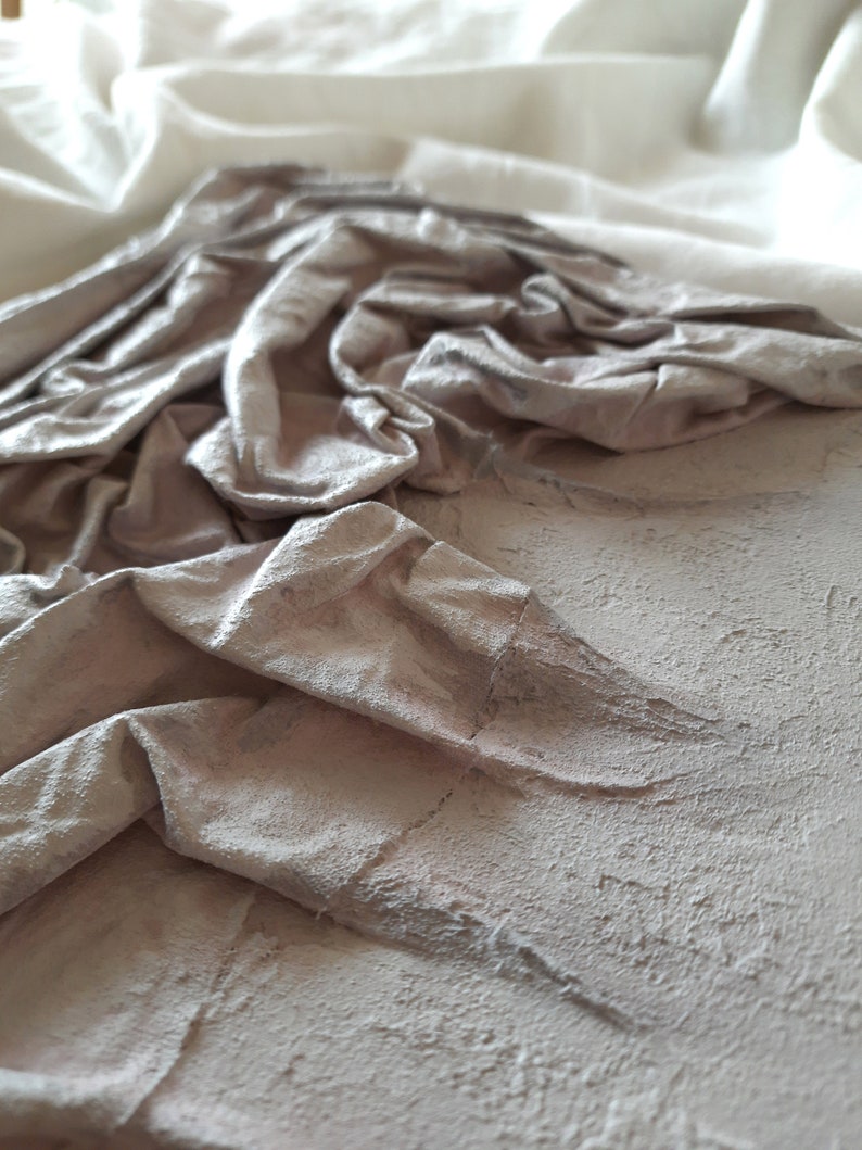 K&ouml;nnte beinhalten: Nahaufnahme einer strukturierten, hellgrauen Oberfl&auml;che mit einem zerknitterten Stoffst&uuml;ck darauf. Der Stoff ist hellgrau und hat eine weiche, knitterige Textur.