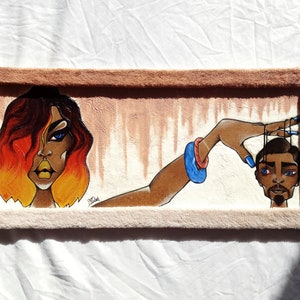 May include: A framed painting featuring a woman with fiery orange hair and a man's head being controlled by strings. The woman is wearing a blue bracelet and has a serious expression. The man's head is brown with a black beard and he has blue eyes.
