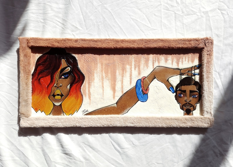 May include: A framed painting featuring a woman with fiery orange hair and a man's head being controlled by strings. The woman is wearing a blue bracelet and has a serious expression. The man's head is brown with a black beard and he has blue eyes.