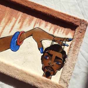 May include: A framed painting depicting a person with blue eyes and a beard. The person is being held by a hand wearing blue and red bracelets. The painting is set against a white background and has a brown frame.