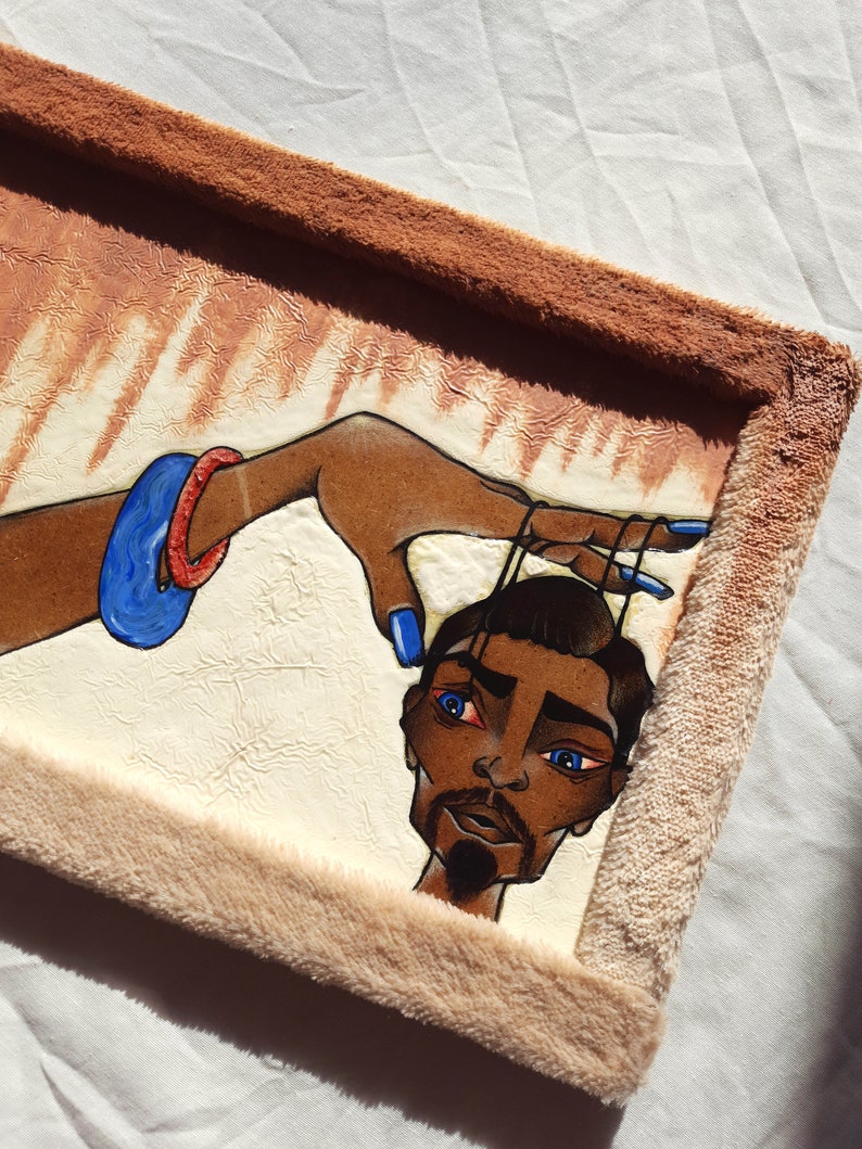 May include: A framed painting depicting a person with blue eyes and a beard. The person is being held by a hand wearing blue and red bracelets. The painting is set against a white background and has a brown frame.