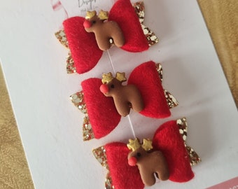 May include: Three red felt hair bows with gold glitter accents and a brown reindeer embellishment with a gold star on its head. The bows are attached to a white card with the text "My Little Love Designs".