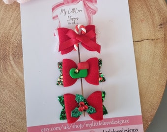 May include: A set of three Christmas-themed hair bows on a white card. The bows are red with festive embellishments, including a candy cane, an elf shoe, and holly. The card has the text "My Little Love Designs".