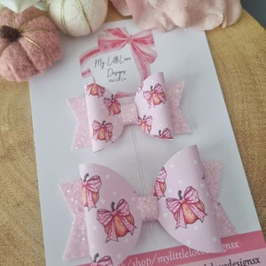 Könnte beinhalten: Zwei rosa Haarschleifen mit einem Kürbismuster. Die Schleifen sind mit Glitzer und Tupfen verziert. Auf der Karte hinter den Schleifen steht "My Little Love Designs" und Social-Media-Informationen.