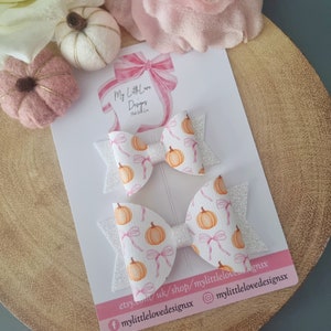 以下が含まれることがあります： キラキラのアクセントとカボチャ柄の白いヘアリボン2つ。リボンは「My Little Love Designs」の文字が入った白いカードにあります。リボンはオレンジ色のカボチャとピンクのリボンで飾られています。