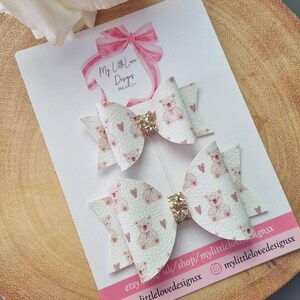 Könnte beinhalten: Zwei weiße Haarbänder mit einem rosafarbenen Teddybär-Druck und goldenen Glitzerakzenten. Die Schleifen befinden sich auf einer weißen Karte mit dem Text "My Little Love Designs" und Social-Media-Handles.