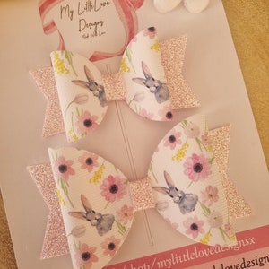 May include: Two pink glitter hair bows with a white floral and bunny print. The bows are on a white card with the text "My Little Love Designs Made With Love" and the text "etsy.com/uk/shop/mylittlelovedesignsx mylittlelovedesignsx @mylittlelovedesign"