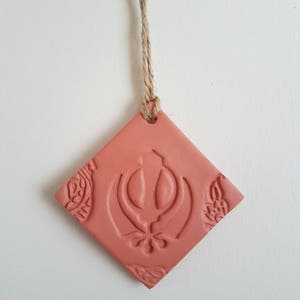 May include: A square, terracotta-coloured ornament with a religious symbol embossed in the centre. The ornament is suspended by a natural fibre cord. Intricate floral patterns are embossed in the corners of the square.