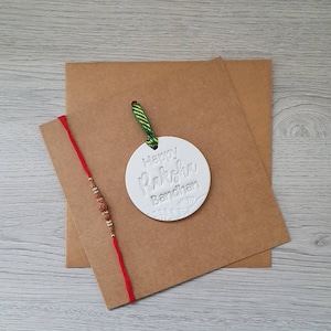 Könnte beinhalten: Eine weiße runde Karte mit dem Text "Happy Raksha Bandhan" in einer dekorativen Schriftart. Die Karte ist an einer braunen Karte mit einer roten Schnur und braunen Perlen befestigt. Die Karte liegt auf einer Holzoberfläche.