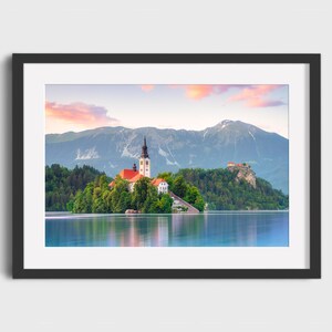 May include: Framed print of a scenic landscape featuring a church on an island in a lake, with mountains in the background. The sky displays a gradient of pink and orange hues. The water reflects the sky and surrounding scenery.