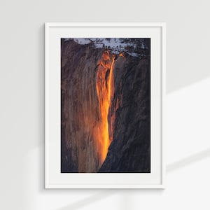May include: A framed photograph of a waterfall illuminated by golden sunlight. The water cascades down a dark, textured rock face, creating a striking contrast of light and shadow. The image is set in a white frame.