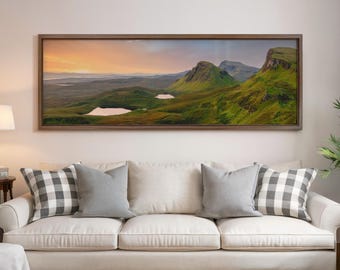 May include: A framed landscape print featuring a scenic view of green hills, two white lakes, and a colorful sunset. The artwork is displayed above a cream-colored sofa with gray and checkered pillows.