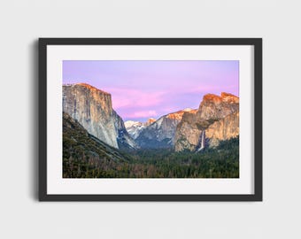 May include: A framed landscape photograph of Yosemite Valley at sunset. The image features towering granite cliffs, a waterfall, and a forest, all under a pink and purple sky. The photograph is in a black frame.