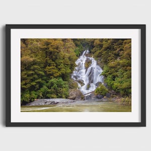 May include: A framed photograph of a waterfall cascading down a rocky cliff, surrounded by lush green and golden-brown trees. The water flows into a river at the base of the falls, creating a serene landscape.
