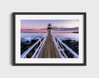 May include: A framed print of a lighthouse at dusk. The wooden pier leads to the white and black lighthouse, with a calm ocean and colorful sky in the background. The sky transitions from pink to purple.