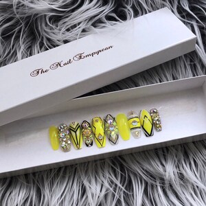 May include: A set of ten yellow press-on nails with black and white geometric designs and rhinestones. The nails are in a white box with the text "The Nail Empyrean".
