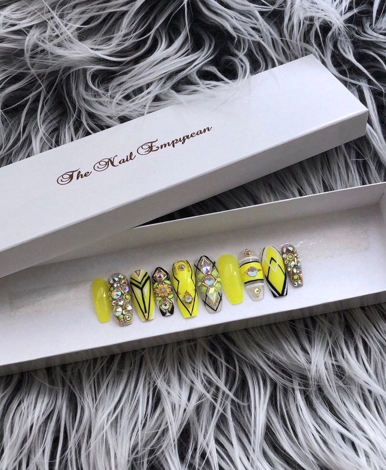 May include: A set of ten yellow press-on nails with black and white geometric designs and rhinestones. The nails are in a white box with the text "The Nail Empyrean".