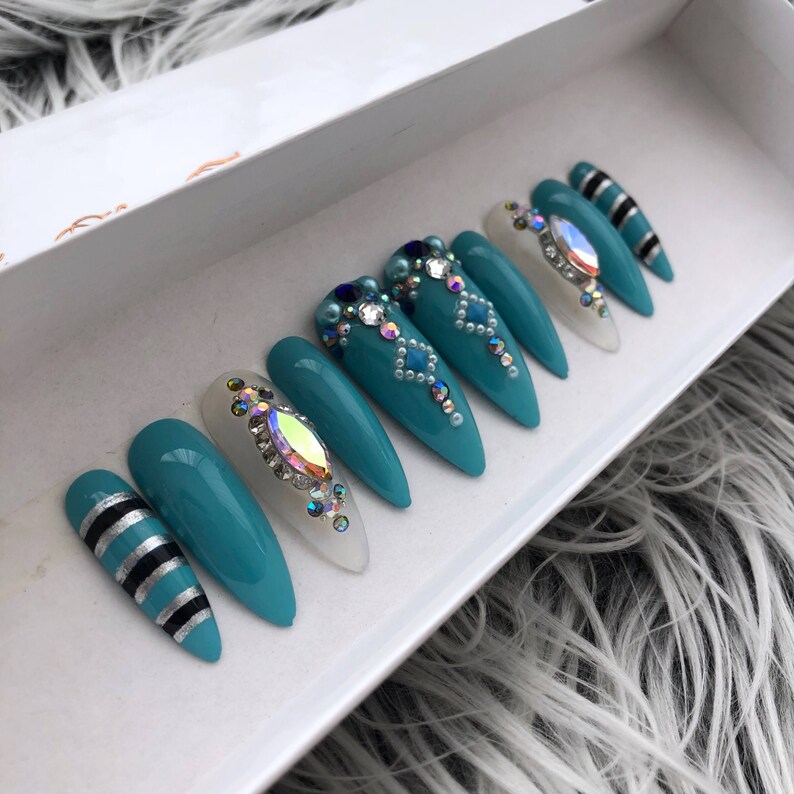 May include: Set of teal blue stiletto false nails with white tips and rhinestones. Some nails have a geometric design with white and silver accents.