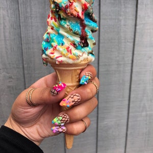 May include: A hand holding a colorful ice cream cone with sprinkles. The nails are painted with a design that looks like ice cream cones with sprinkles.