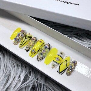 May include: A set of ten yellow press-on nails with black and white geometric designs and rhinestones. The nails are in a white box with the text "The Nail Empyrean".
